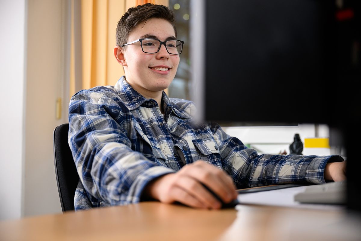 Ein lächelnder Junge in einem karierten Hemd sitzt an einem Tisch, hält eine Computermaus in seiner Hand und schaut auf einen Bildschirm. 